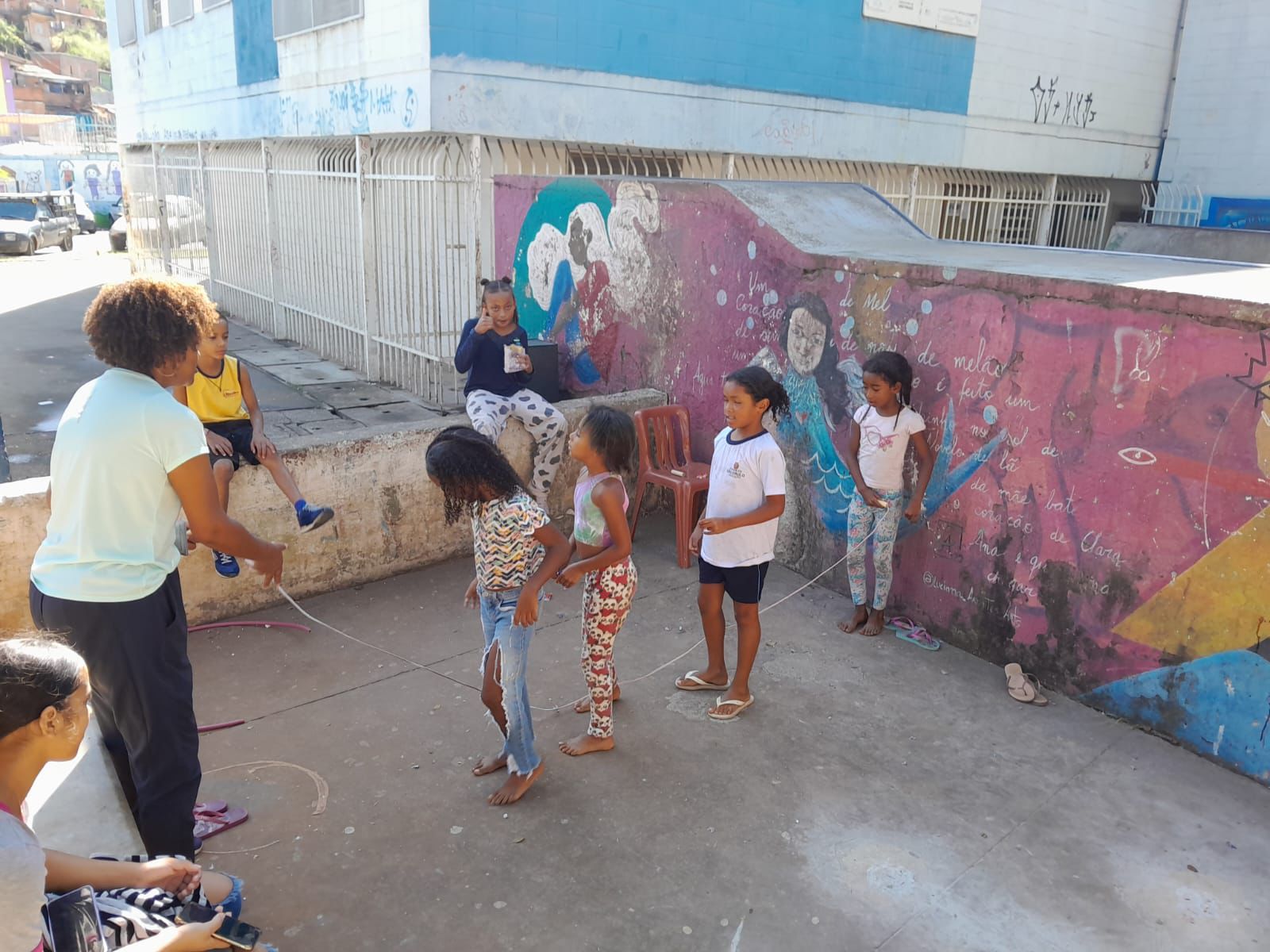 Fotografia com crianças enfileiradas brincando de pular corda, com roupas coloridas. Muros com desenhos coloridos.